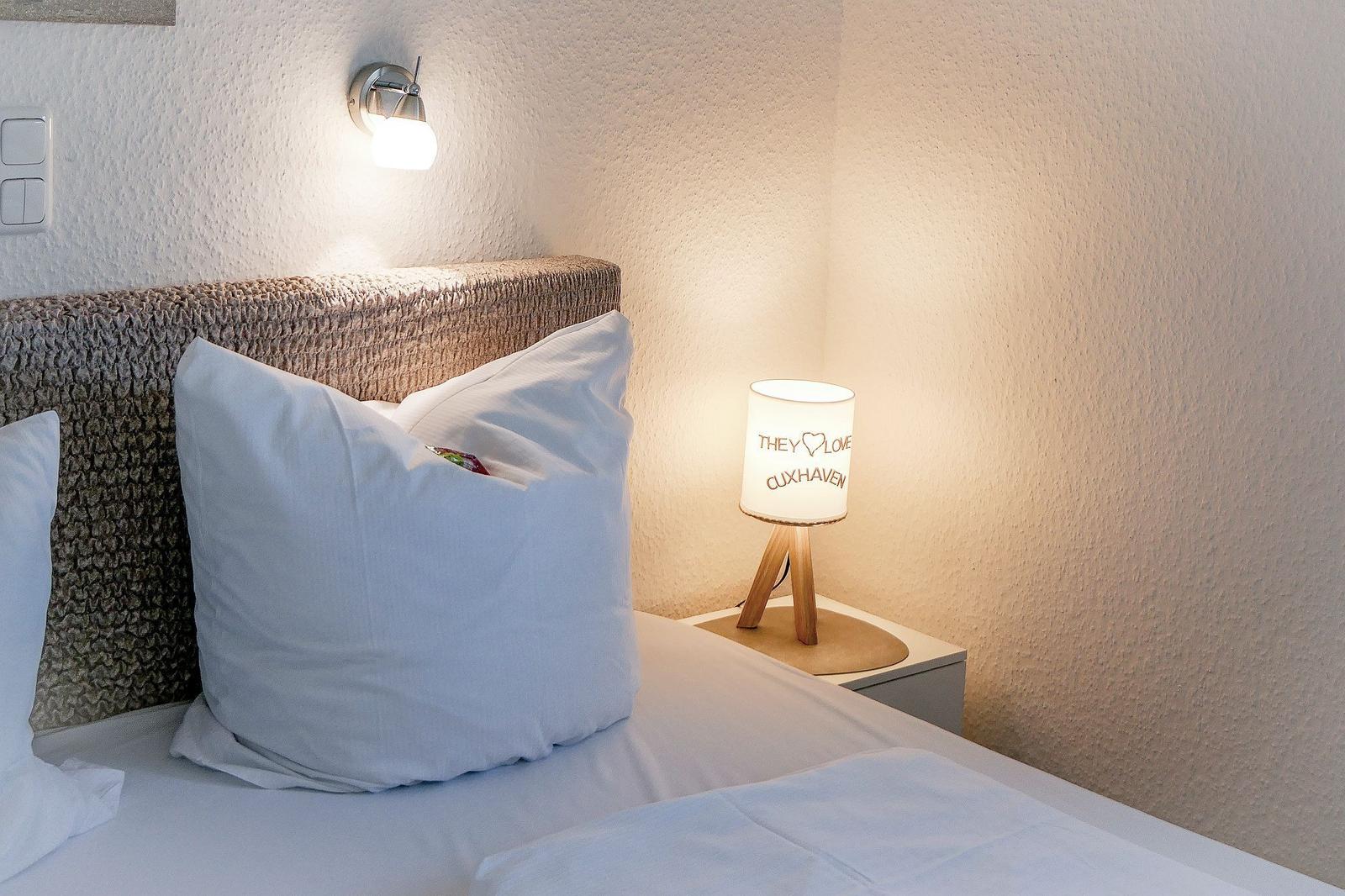 Bettkopfteil mit Kissen, Nachttisch mit Lampe und Wandlampe.