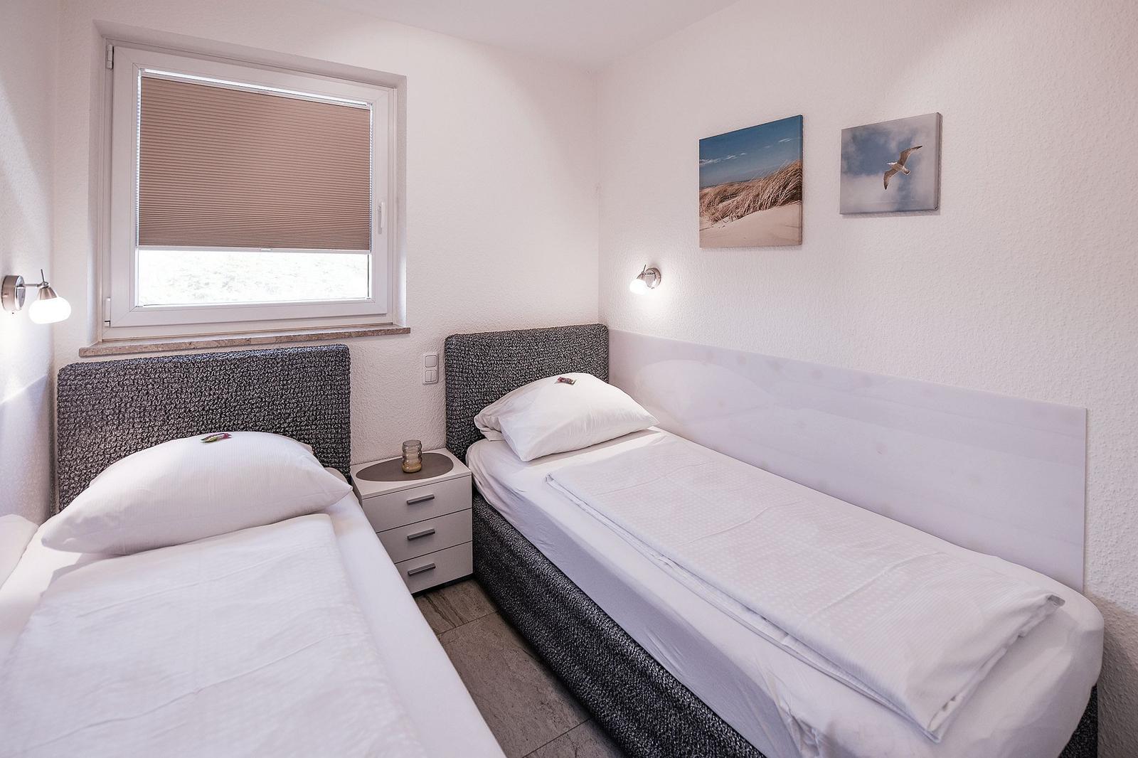 Zwei Einzelbetten mit weißen Bettwäschen, Nachttisch und Fenster mit Jalousie.