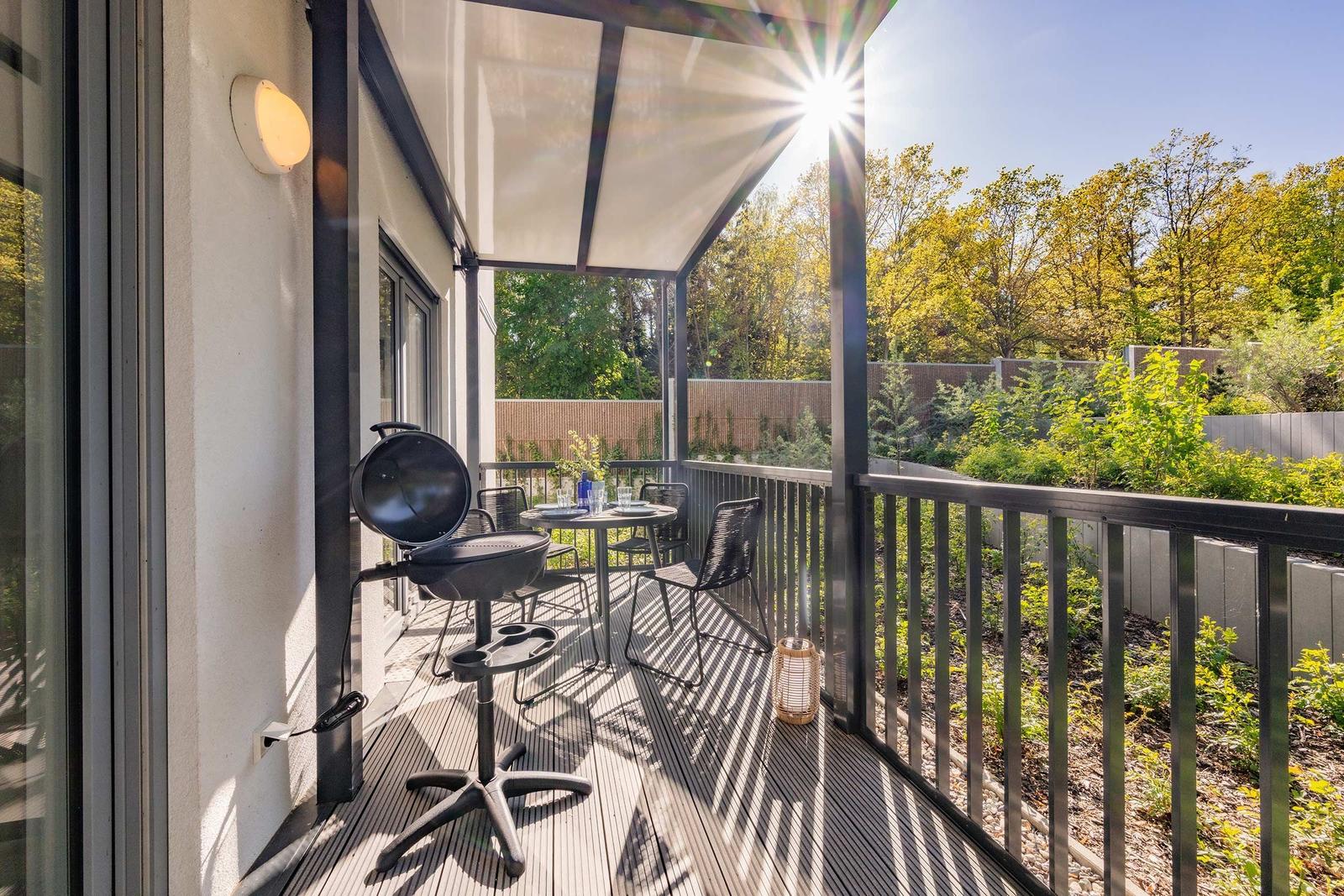 Terrasse mit Grill, Tisch und Stühlen. Sonne scheint durch den Schirm. Grünflächen im Hintergrund.