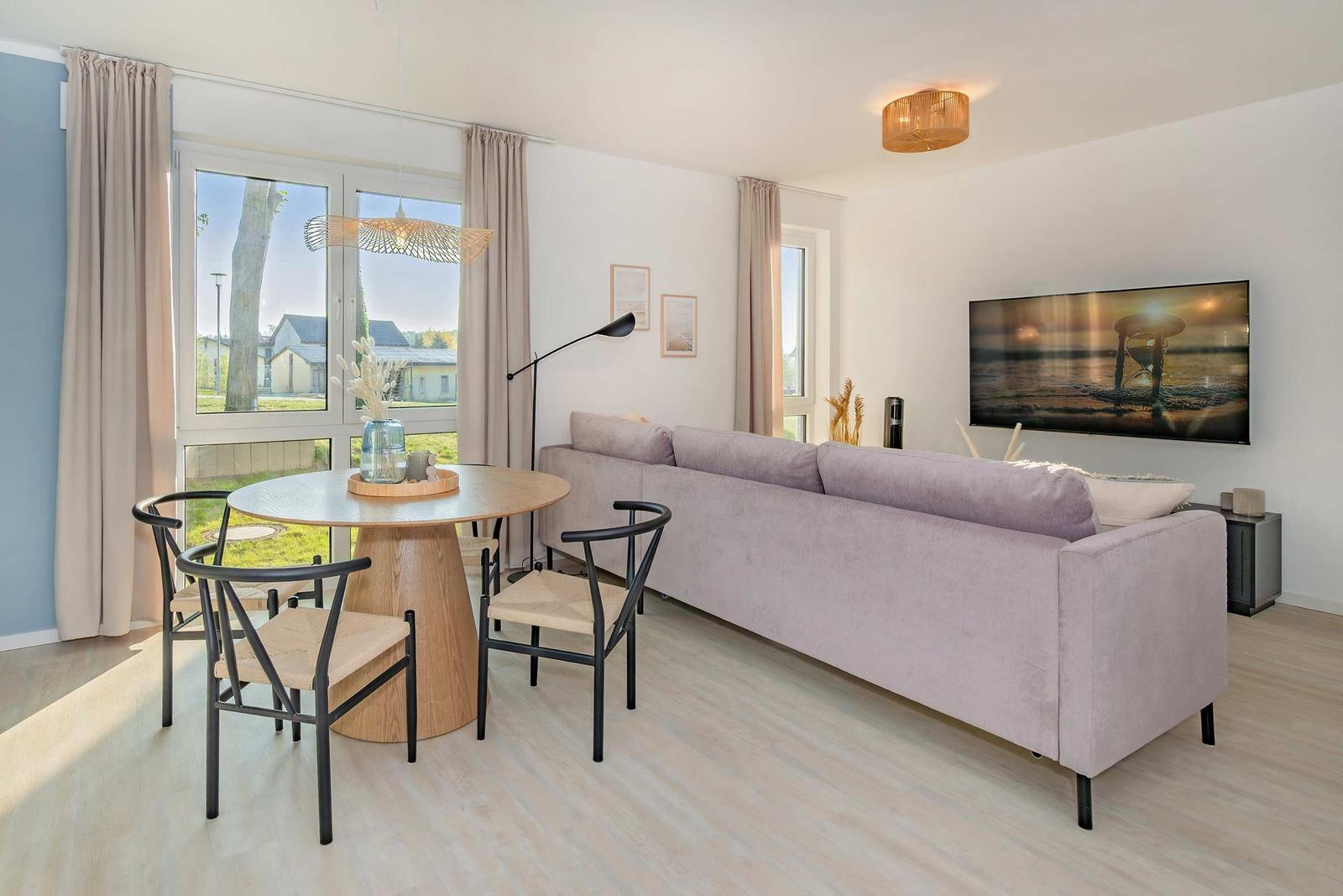 Wohn- und Essbereich mit Sofa, Esstisch und TV. Große Fenster mit Blick nach draußen.