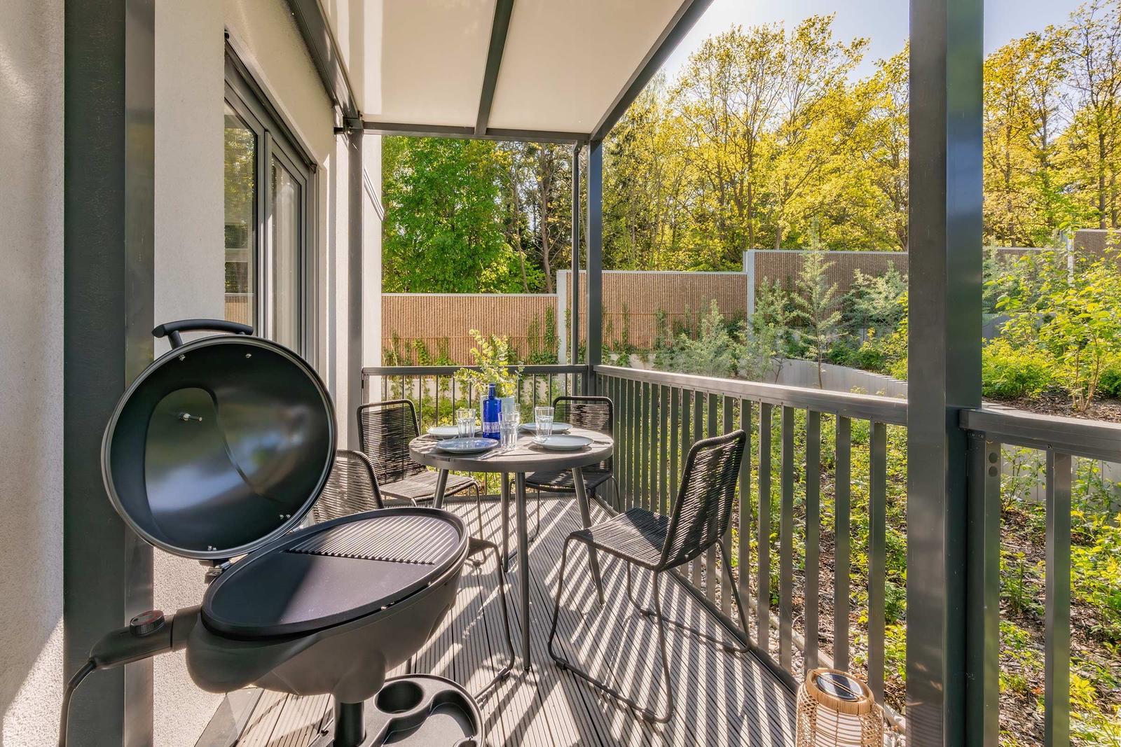 Terrasse mit Grill, Tisch und Stühlen. Grüne Umgebung mit Bäumen und Zaun.