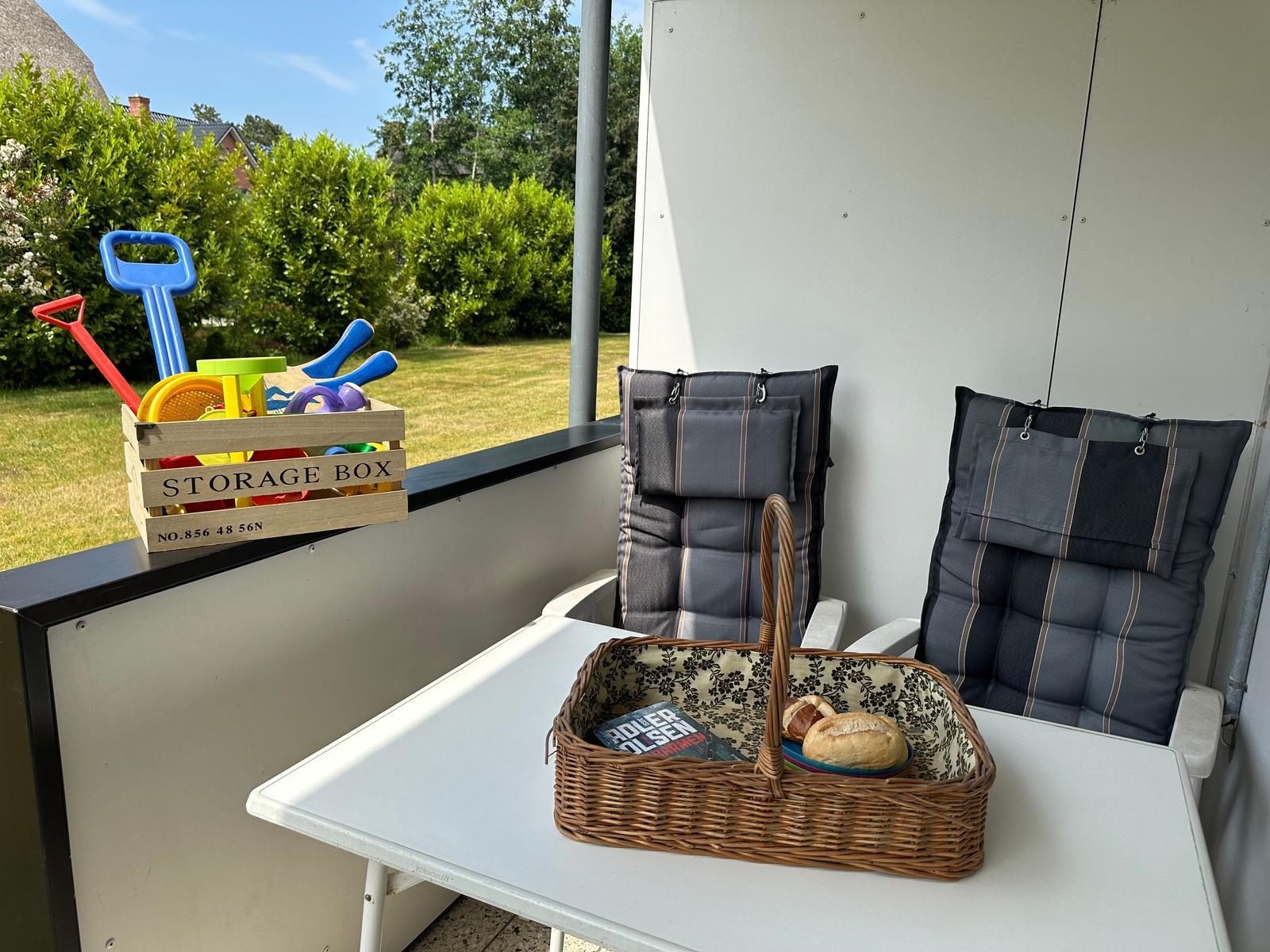Terrasse mit Tisch, zwei Stühlen und Korb mit Brot. Spielzeugkiste auf der Brüstung.