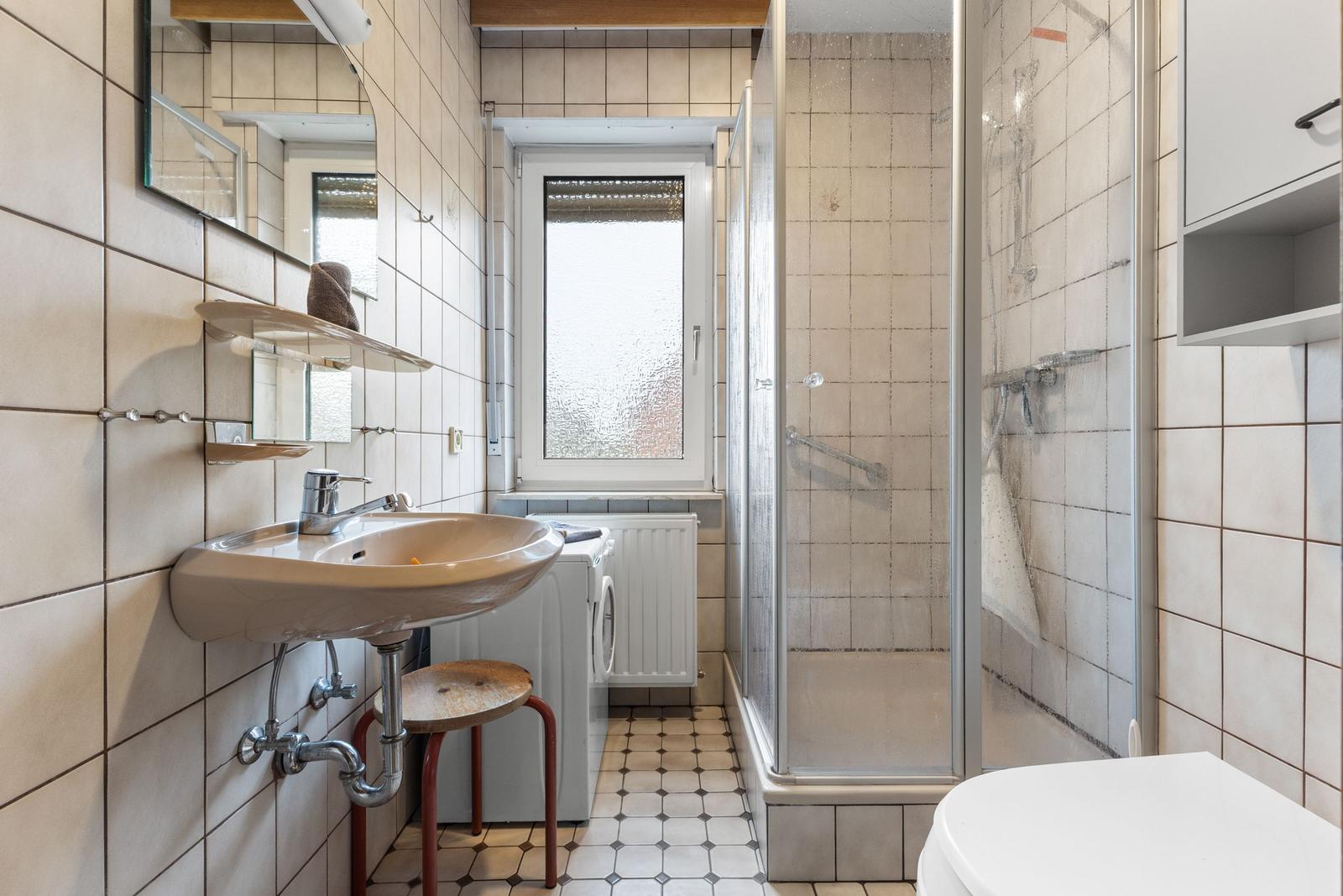 Badezimmer mit Dusche, Waschbecken und Toilette. Fliesenwand und Fenster sichtbar.