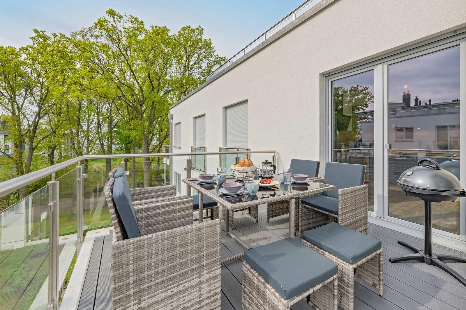 Terrasse mit Sitzgruppe, Tisch und Grill. Blick auf Bäume und Gebäude.