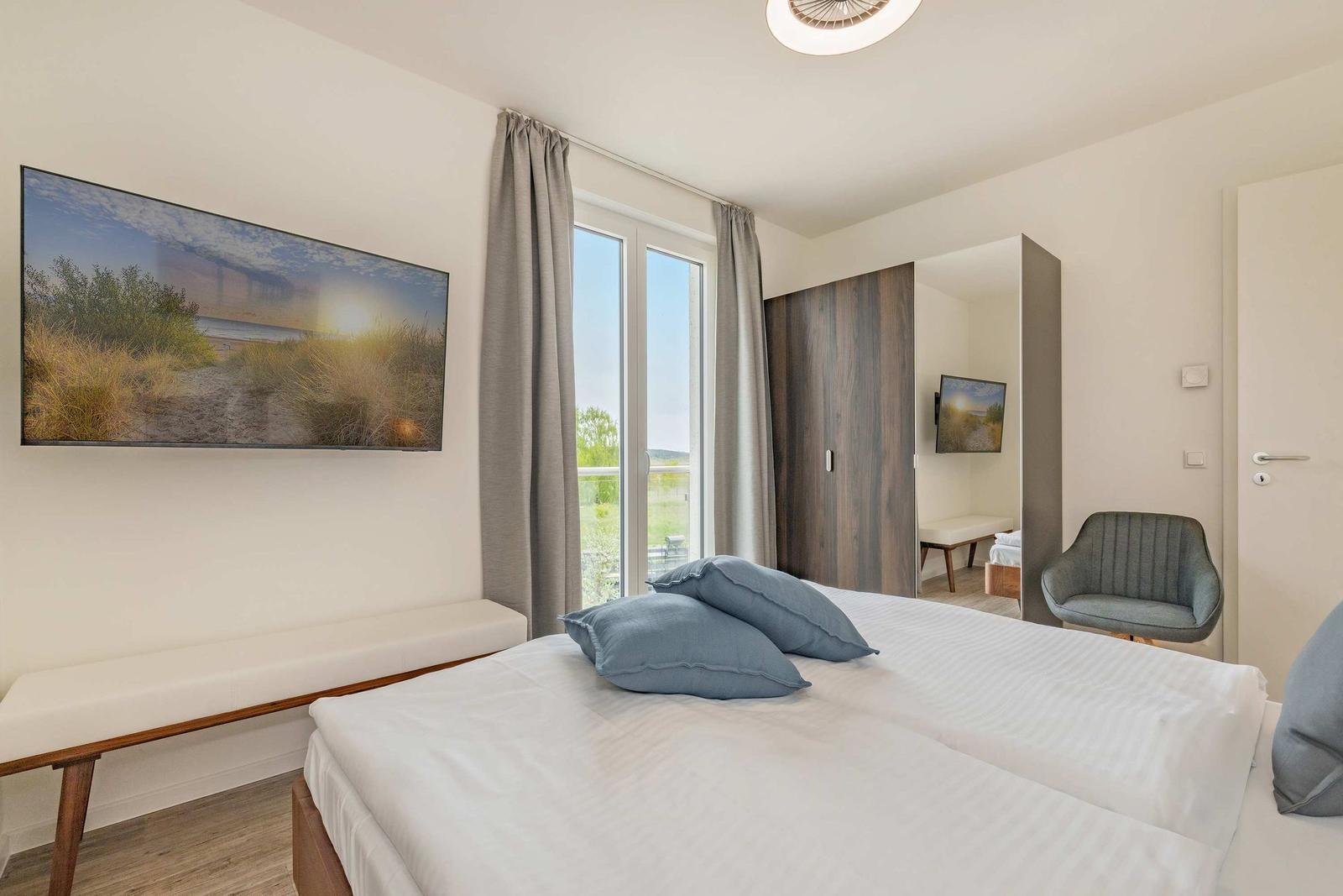 Schlafzimmer mit Bett, Fenster und Wandbild. Sicht auf Balkon. Sessel und Kleiderschrank.