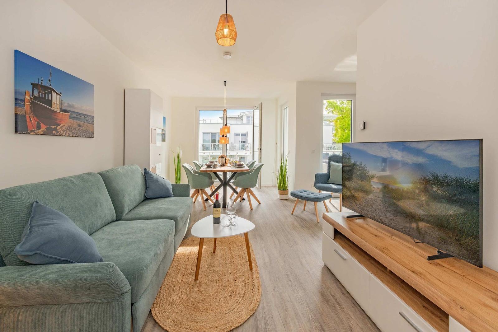 Wohn- und Essbereich mit Sofa, TV und Tisch. Fenster zum Balkon.