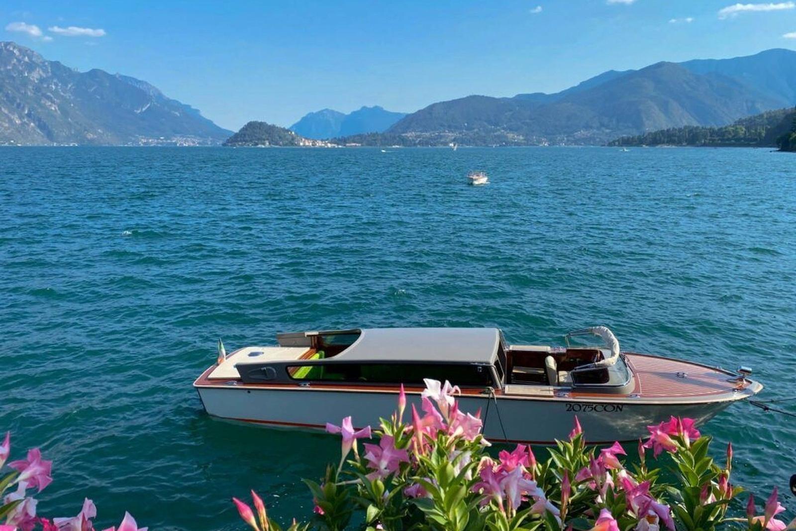 Ein Boot liegt vor einer Bucht mit Bergen im Hintergrund und rosa Blumen im Vordergrund.