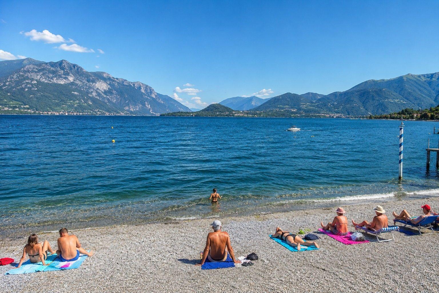 Strand mit Steinen, Menschen baden und entspannen, Berge im Hintergrund, Boot auf dem Wasser.