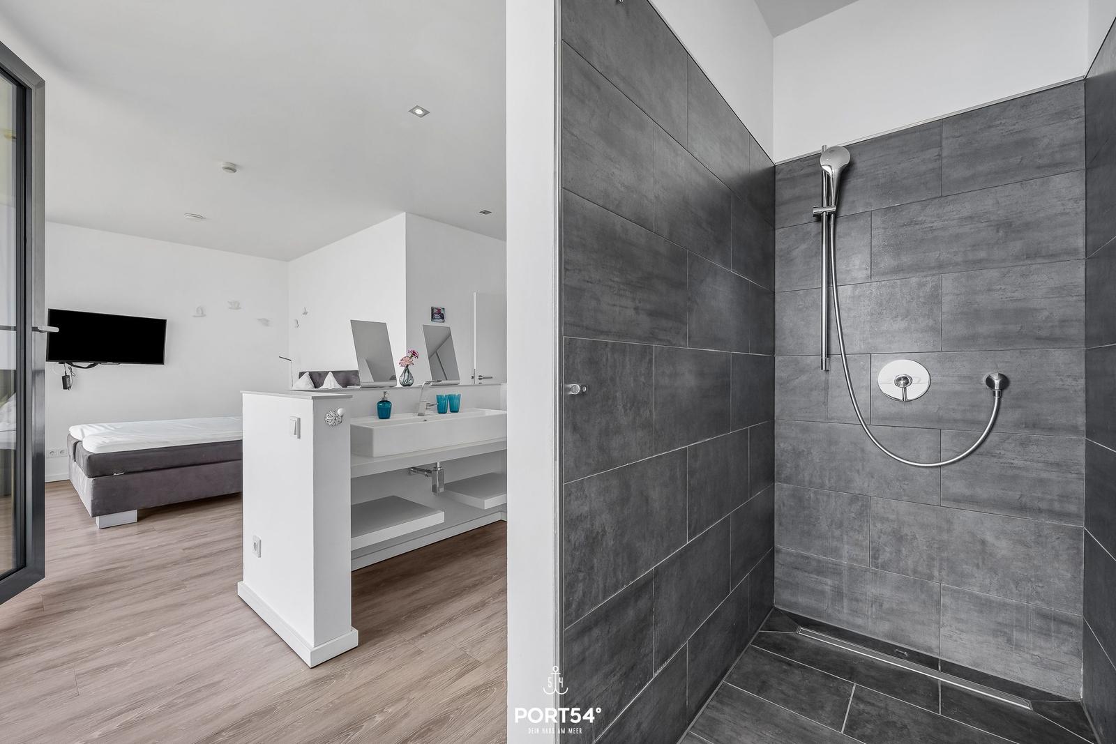 Dusche mit grauen Fliesen, Waschbecken und Schlafzimmer im Hintergrund.