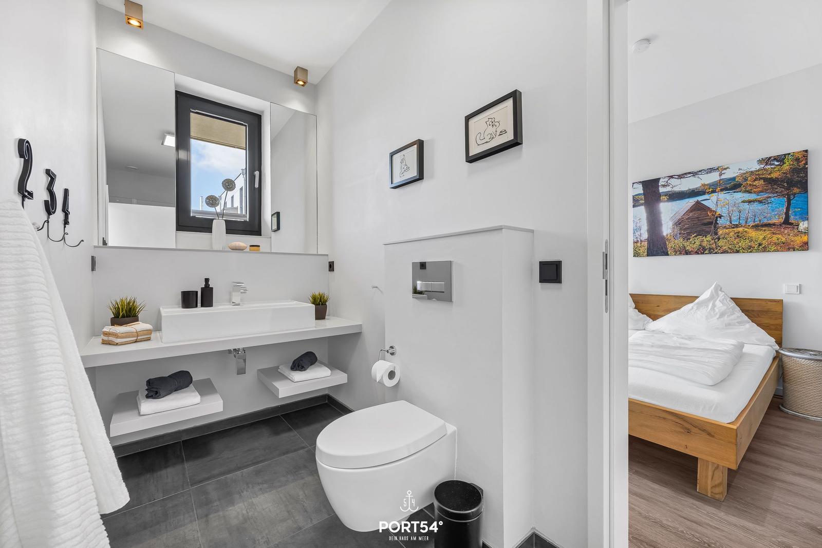 Badezimmer mit Waschbecken, Toilette und Fenster. Nebenraum mit Bett und Wandbild.