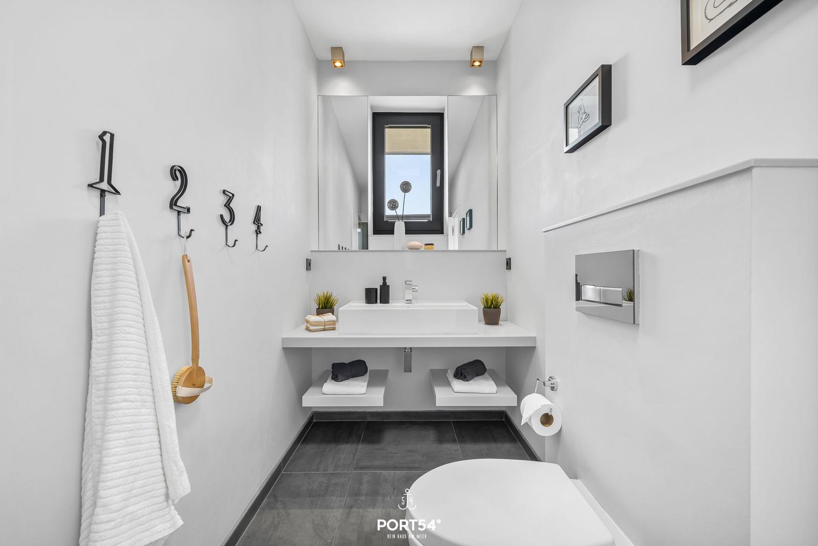Badezimmer mit Waschtisch, Spiegel und Toilette. An der Wand hängen Handtücher und ein Bürstenhalter.