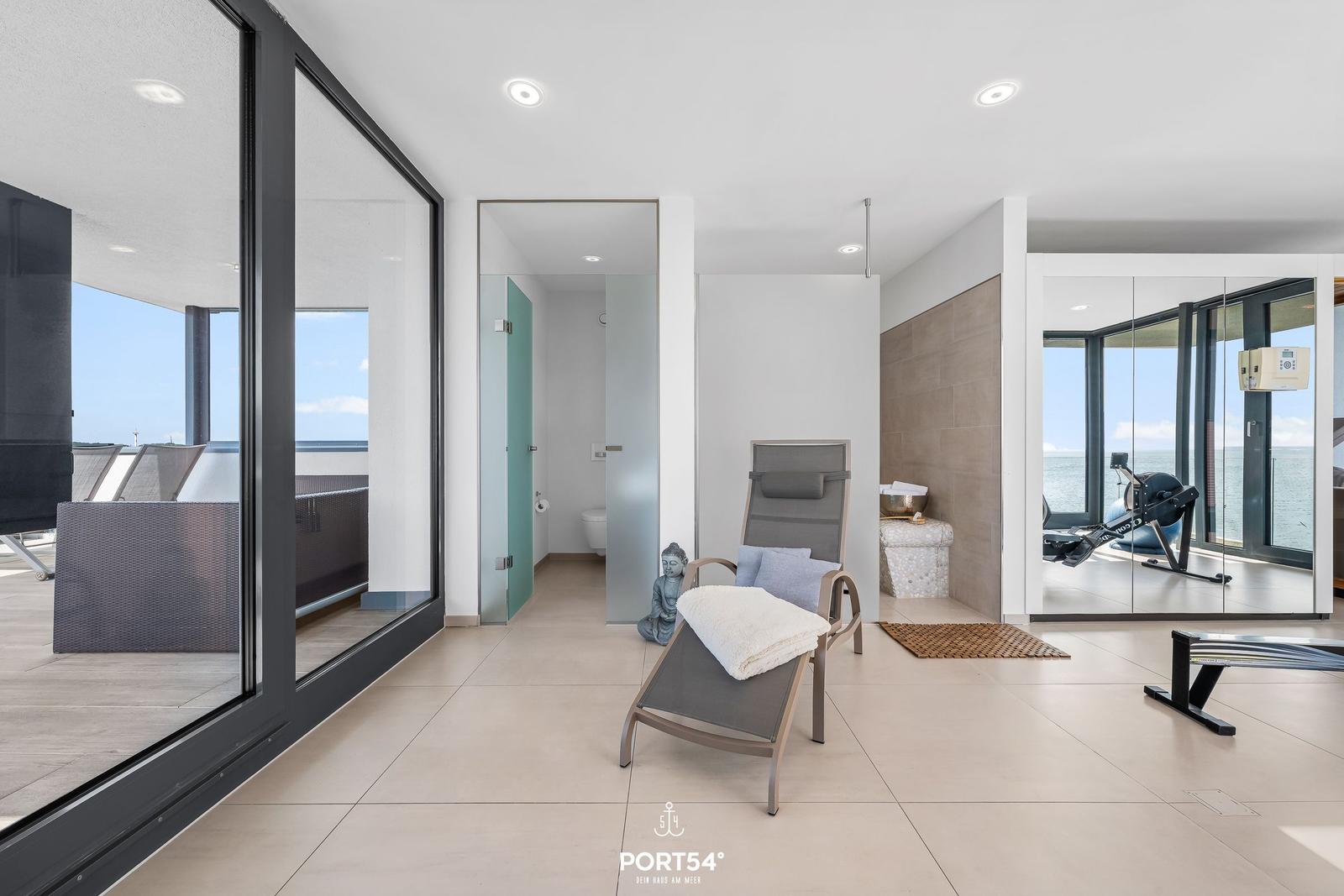 Wohnbereich mit Blick aufs Meer, Liege, Fitnessgeräte und Badzimmer.