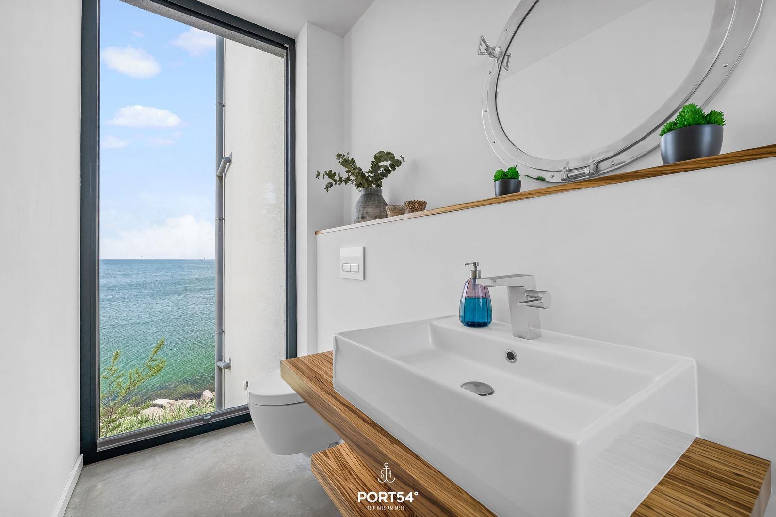 Badezimmer mit Blick aufs Meer, Waschbecken auf Holz, runder Spiegel, Pflanzen und Toilette.