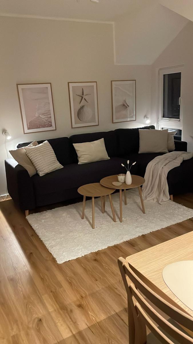 Schwarzes Sofa mit Kissen, zwei runde Holztische und weißer Teppich auf Holzboden.