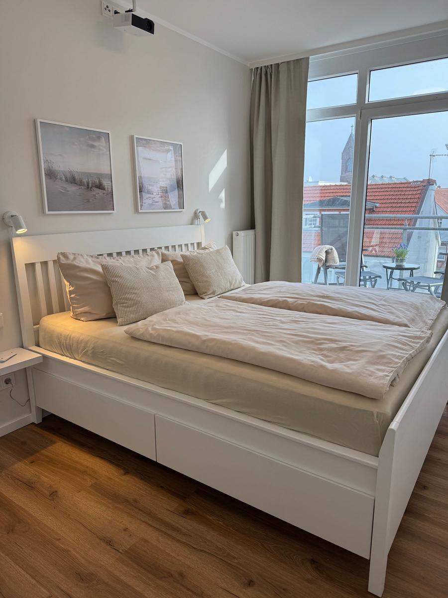 Schlafzimmer mit weißem Bett, Holzboden und Blick auf Balkon.