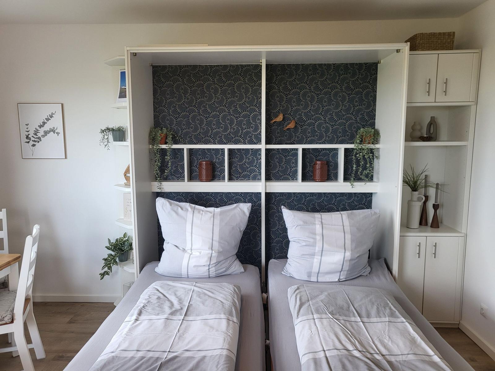 Two beds with white pillows and bedding. Behind the beds is a white wardrobe with dark wallpaper.
