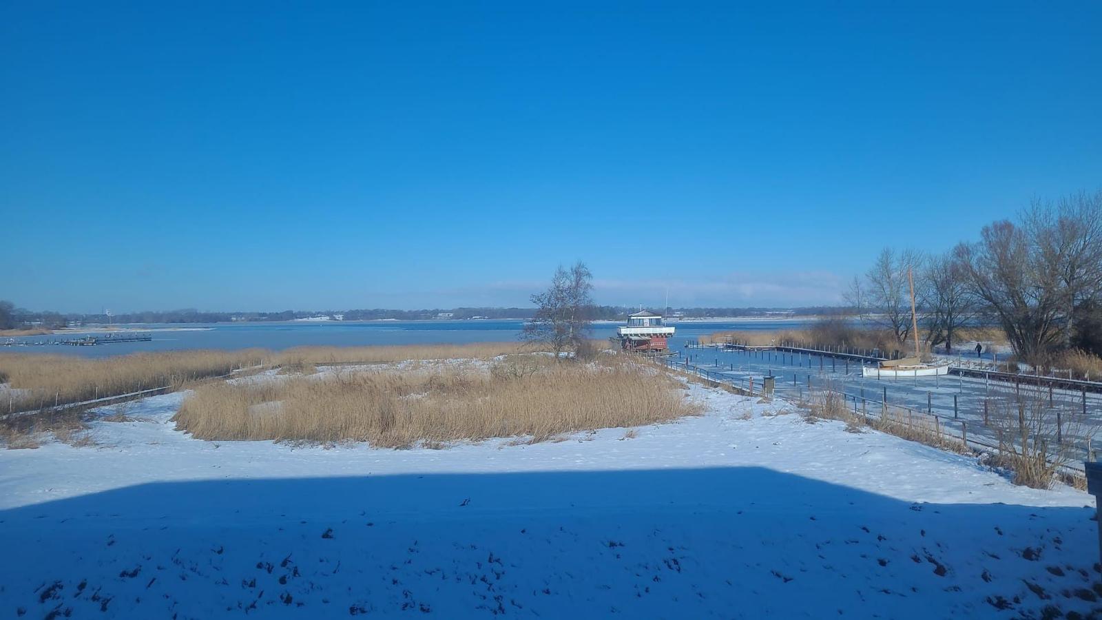 Schneebedeckter Uferbereich mit Blick auf einen See und eine kleine Holzstruktur.