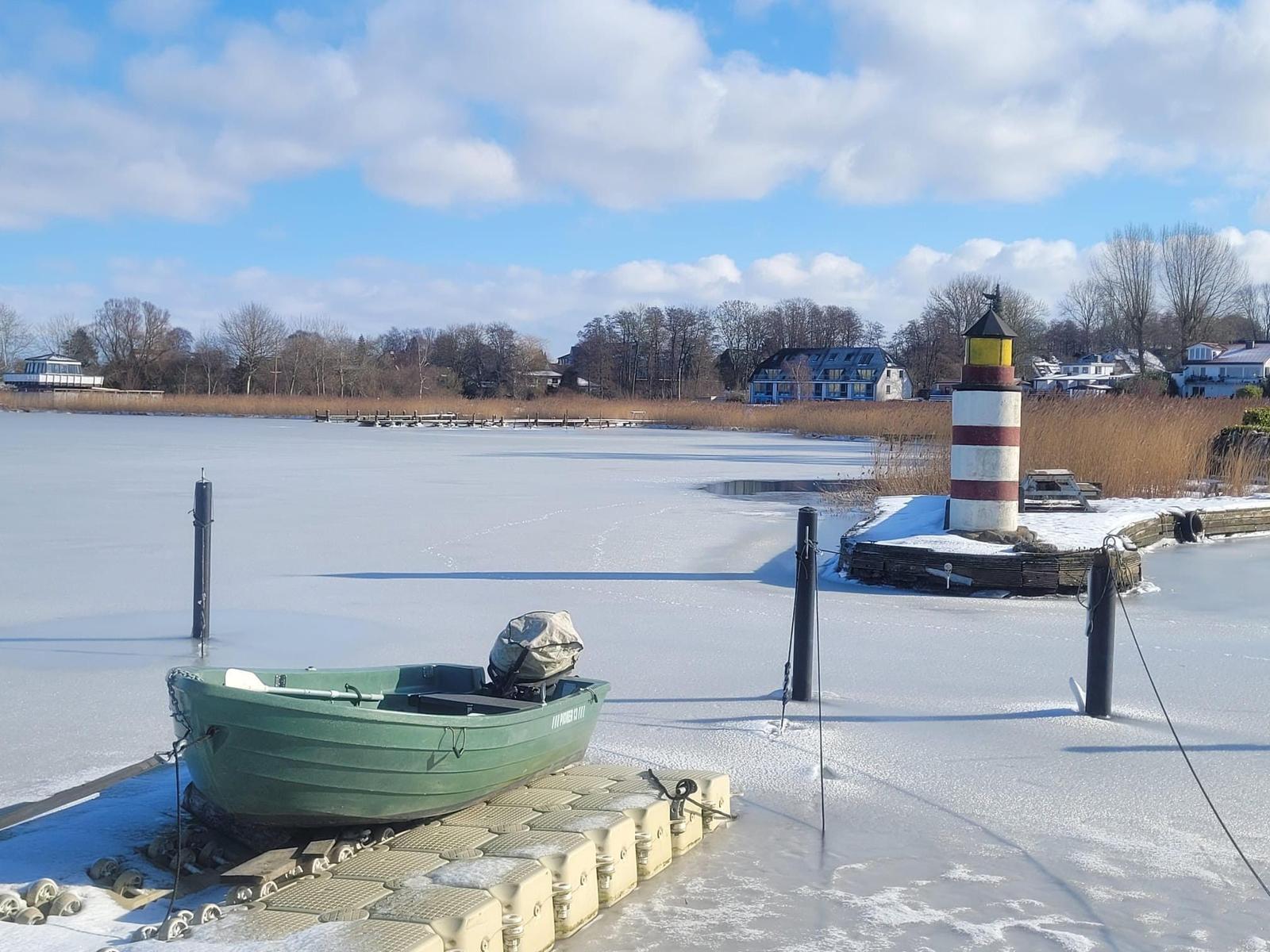 Ein grünes Boot liegt auf einer Eisfläche neben einem Leuchtturm. Im Hintergrund sind Häuser und Bäume zu sehen.