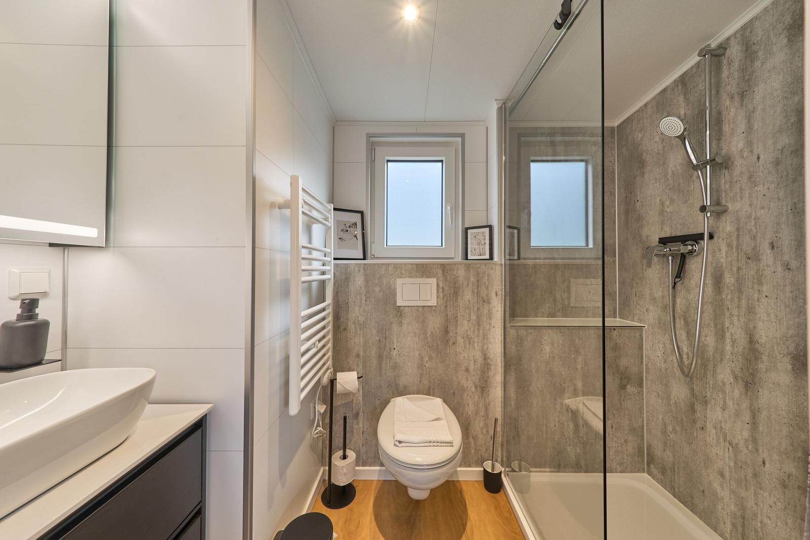 Badezimmer mit Dusche, Toilette und Waschbecken. Holzboden und Betonwand.