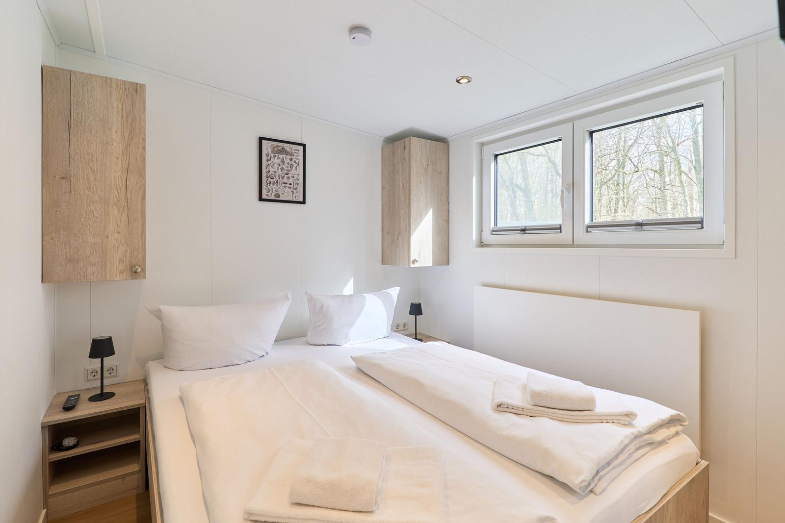 Schlafzimmer mit Doppelbett, zwei Nachttischen und Fenster mit Blick auf Wald.