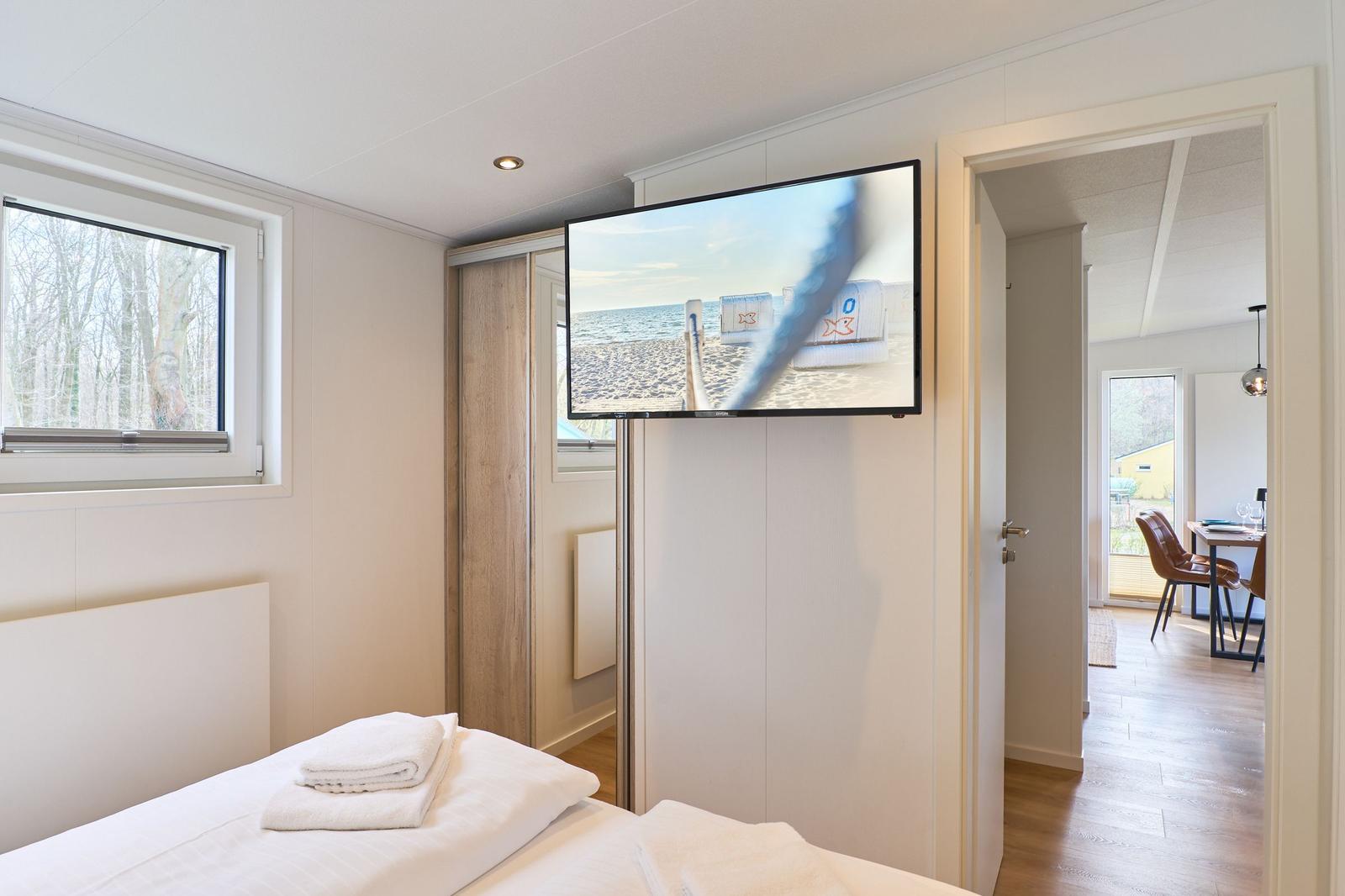Schlafzimmer mit Bett, TV und Fenster. Durchgang zu Wohnbereich mit Tisch und Stühlen.