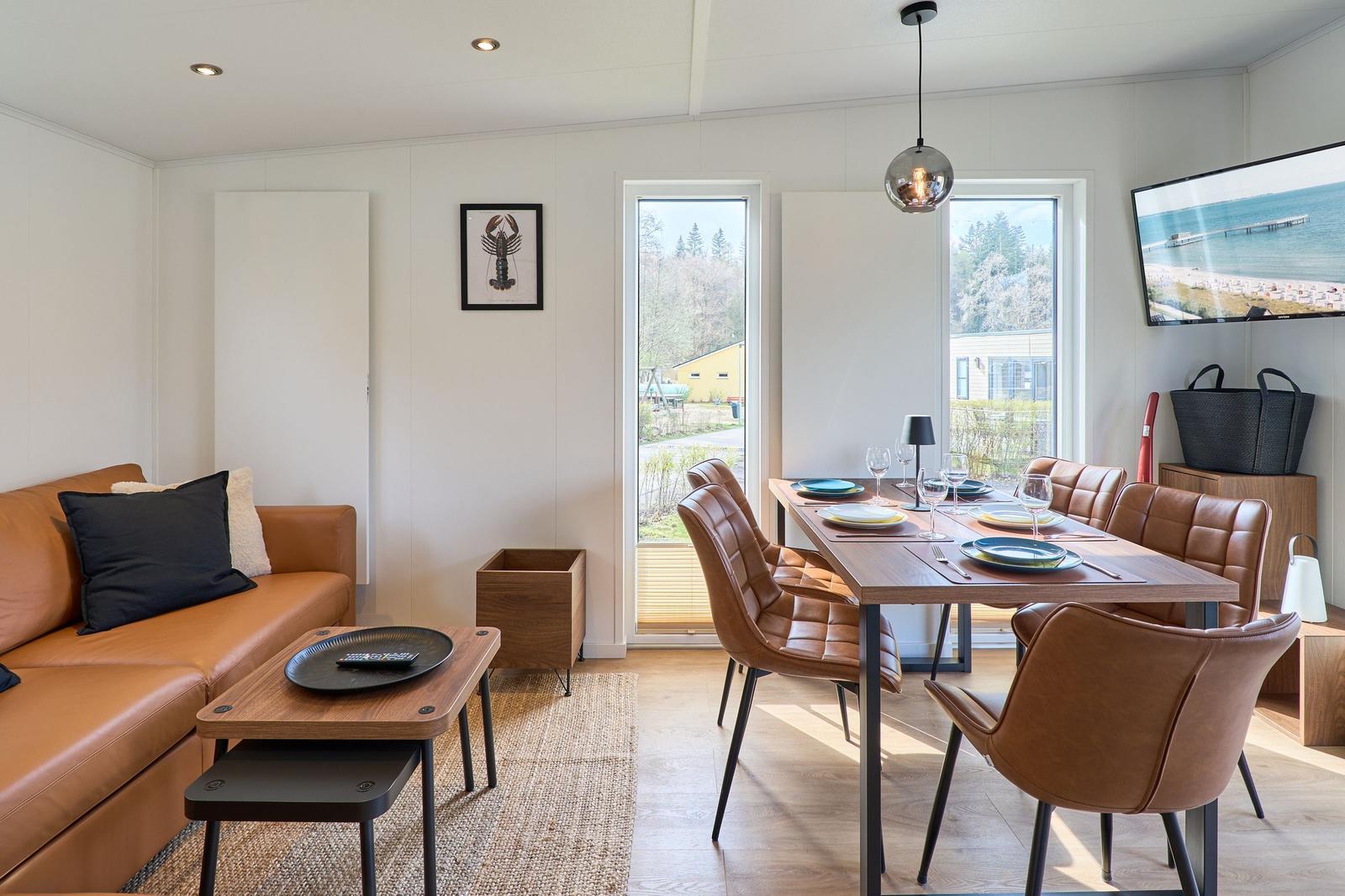 Wohn- und Essbereich mit braunem Sofa, Esstisch und Stühlen. Große Fenster und Wandbild.