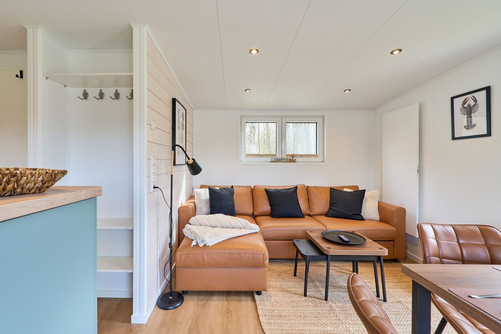 Wohnzimmer mit braunem Sofa, Holztisch und Fenster. Küchenbereich mit blauer Arbeitsplatte.