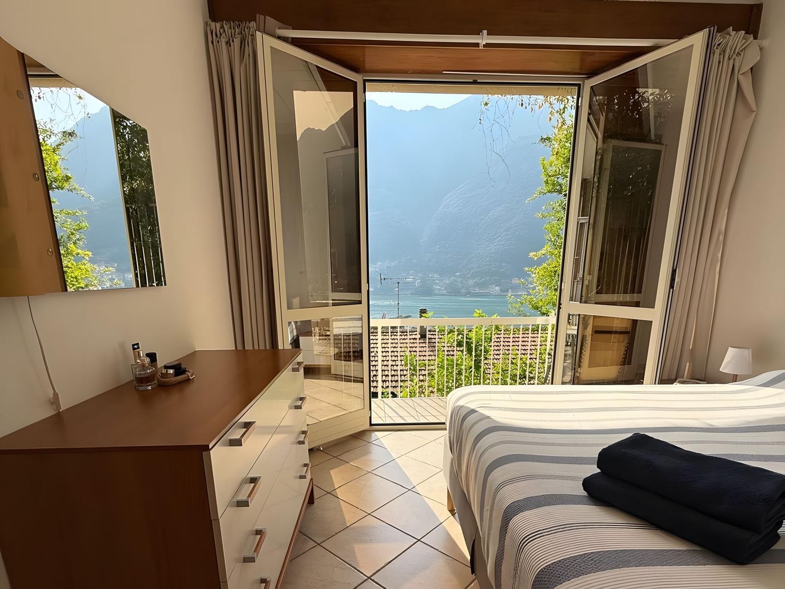 Schlafzimmer mit Balkonblick auf See und Berge. Bett, Schrank und Fenster sichtbar.