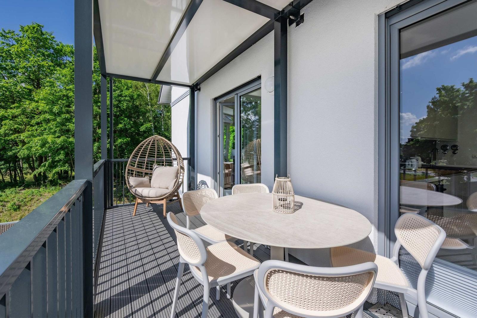 Terrasse mit Esstisch, Stühlen und Hängesessel. Blick auf grüne Bäume. Glasfront zum Innenraum.