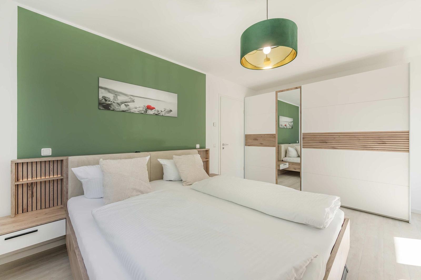Schlafzimmer mit Bett, grüner Wand und Schrank. Deckenleuchte und Bild an der Wand.