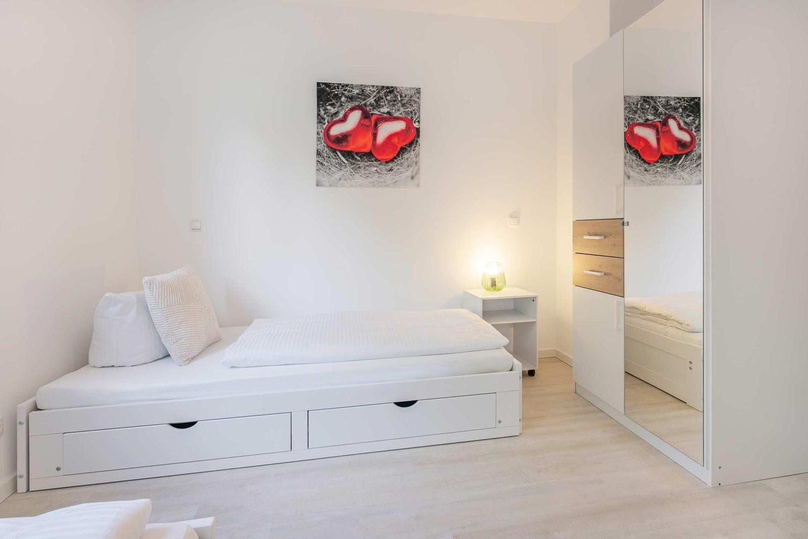 Schlafzimmer mit weißem Bett, Kleiderschrank und Wandbild mit Herzen.