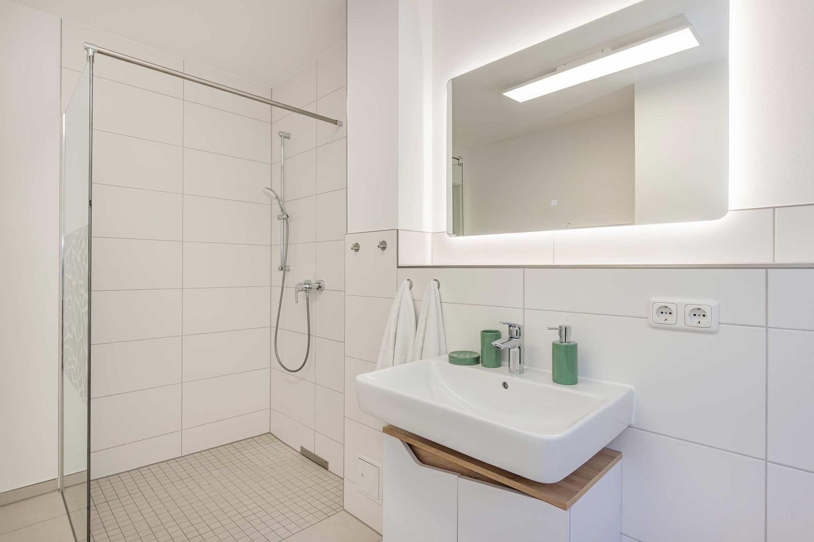 Badezimmer mit Dusche, Waschbecken und beleuchtetem Spiegel.