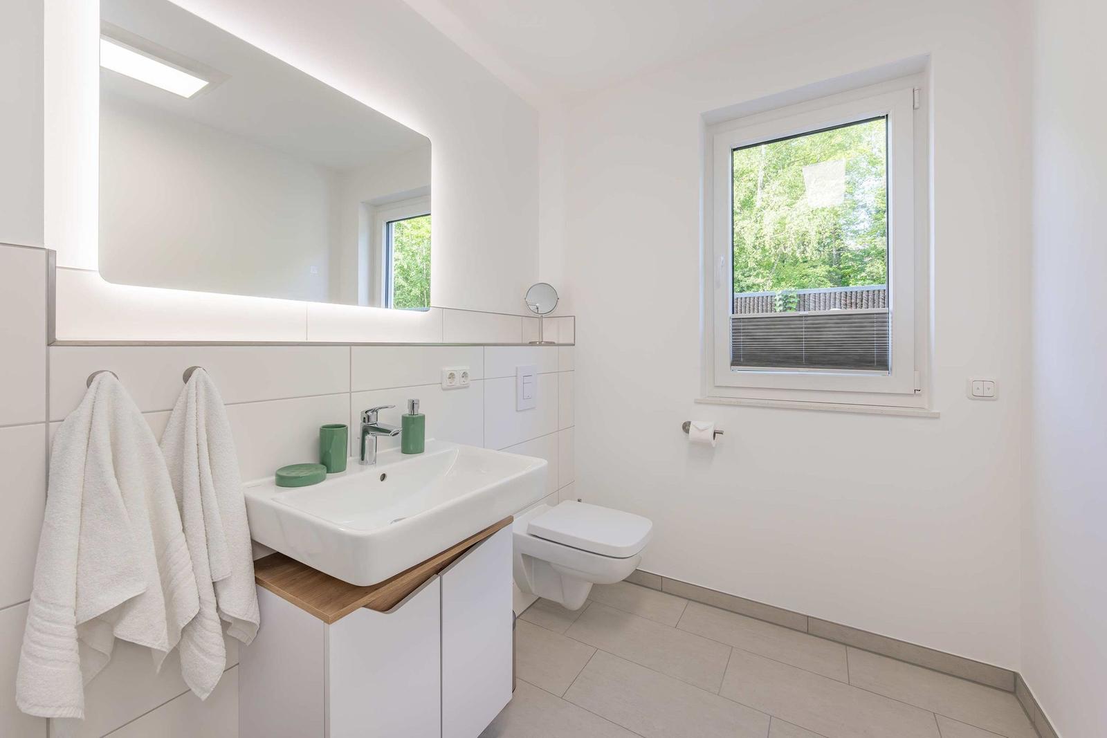 Badezimmer mit Waschbecken, Spiegel, Toilette und Fenster mit Blick auf Grün.