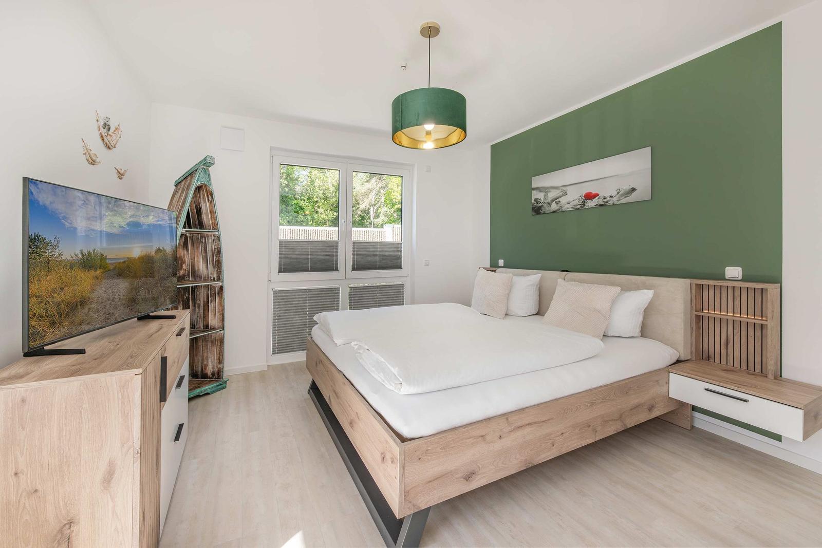 Schlafzimmer mit Bett, Fernseher und Fenster. Wand ist grün. Deckenleuchte und Bilder.