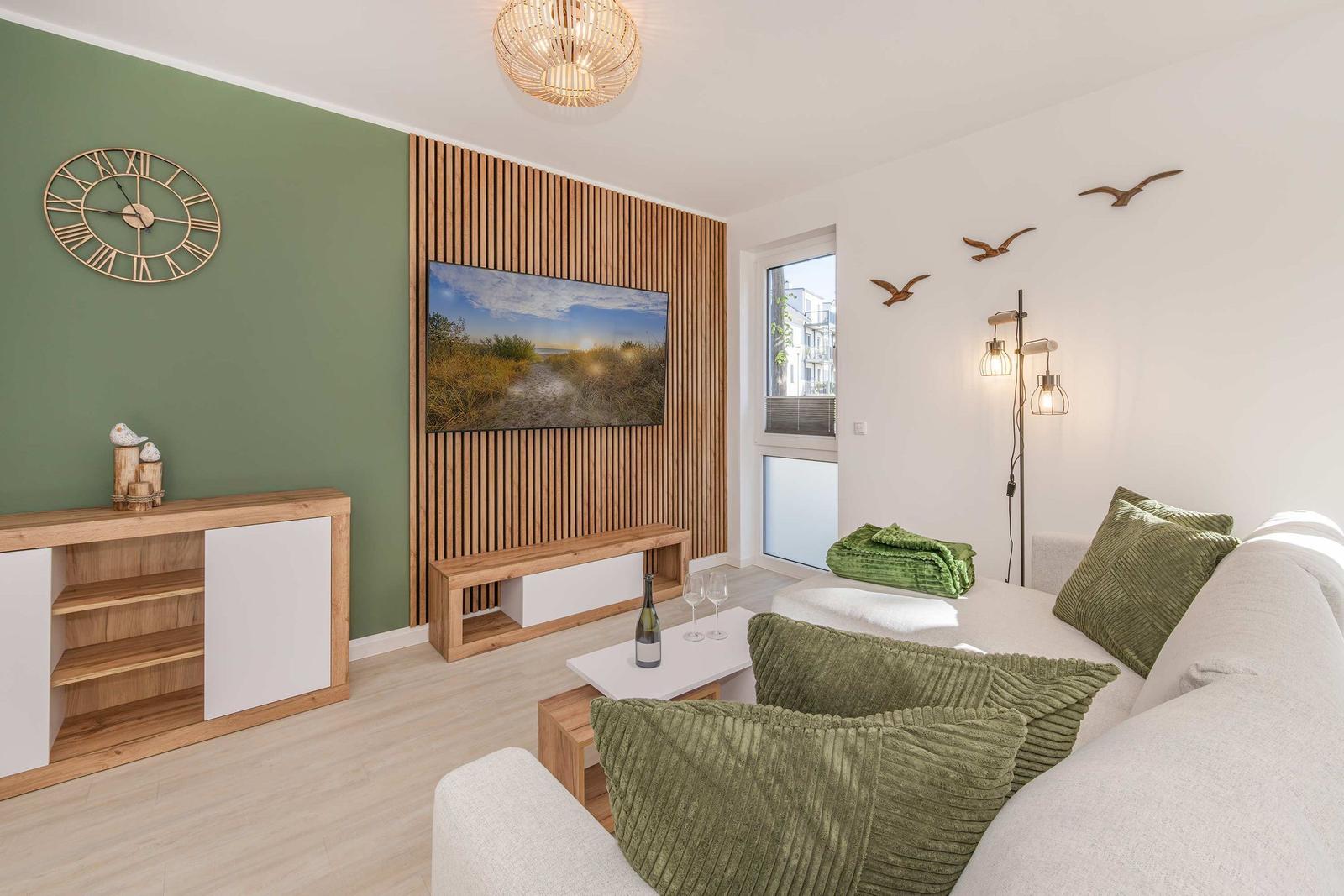 Wohnzimmer mit Sofa, Fernseher und Holzwand. Grüne Kissen und Wanduhr sichtbar.