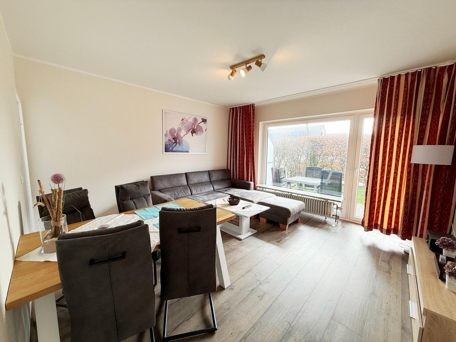 Wohn- und Essbereich mit Sofa, Tisch und Stühlen. Fenster mit Vorhängen zum Garten.