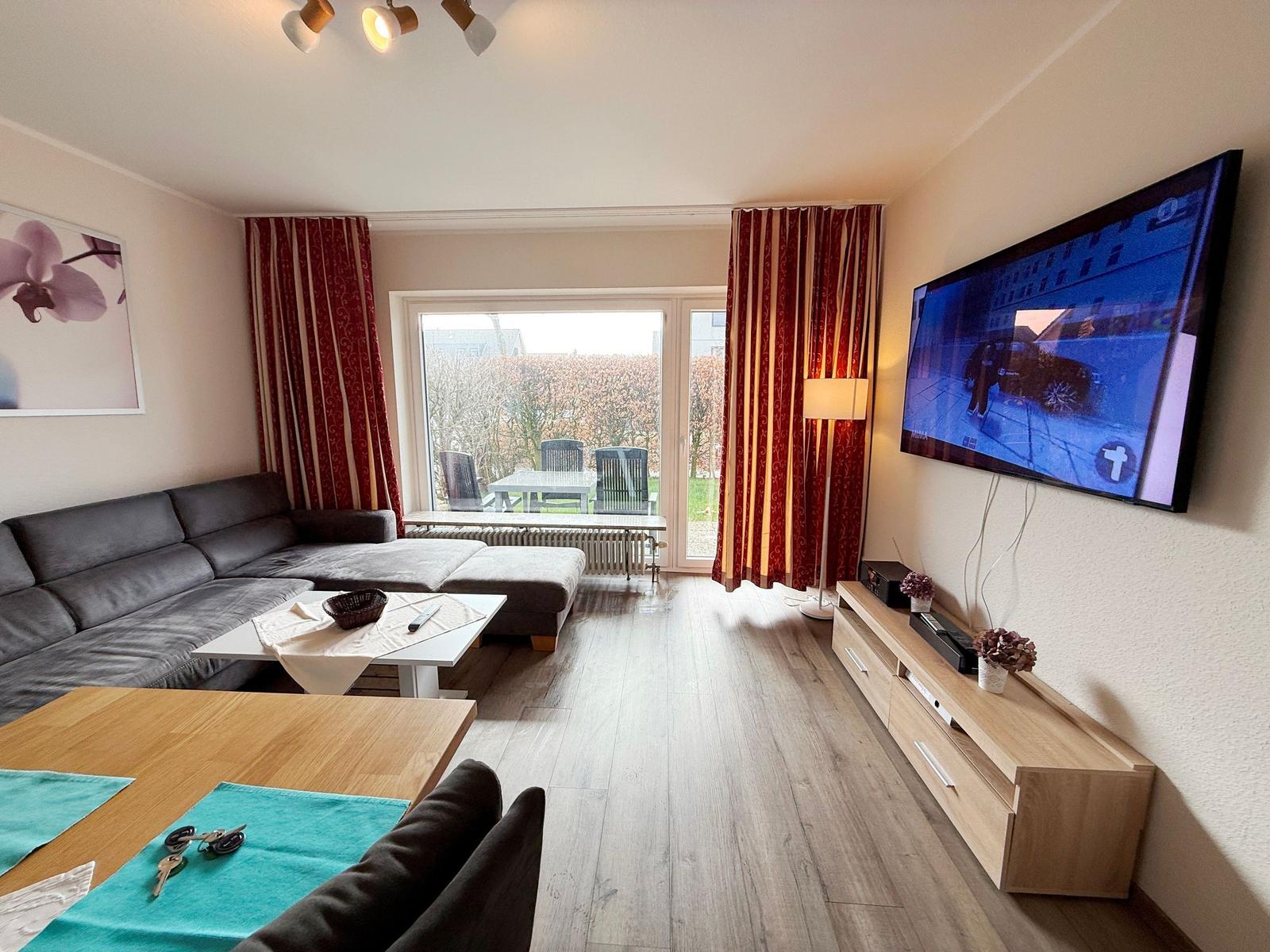 Wohnzimmer mit Sofa, TV und Terrasse. Holzboden und rote Vorhänge.