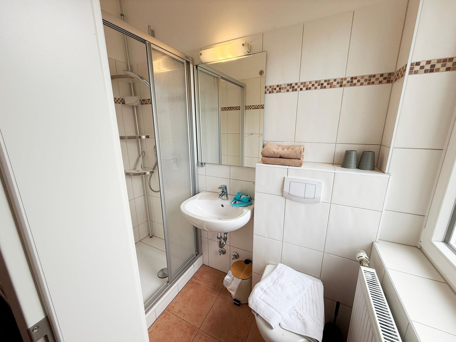 Badezimmer mit Dusche, Waschbecken und Toilette. Weiße Fliesen und braune Bodenfliesen.