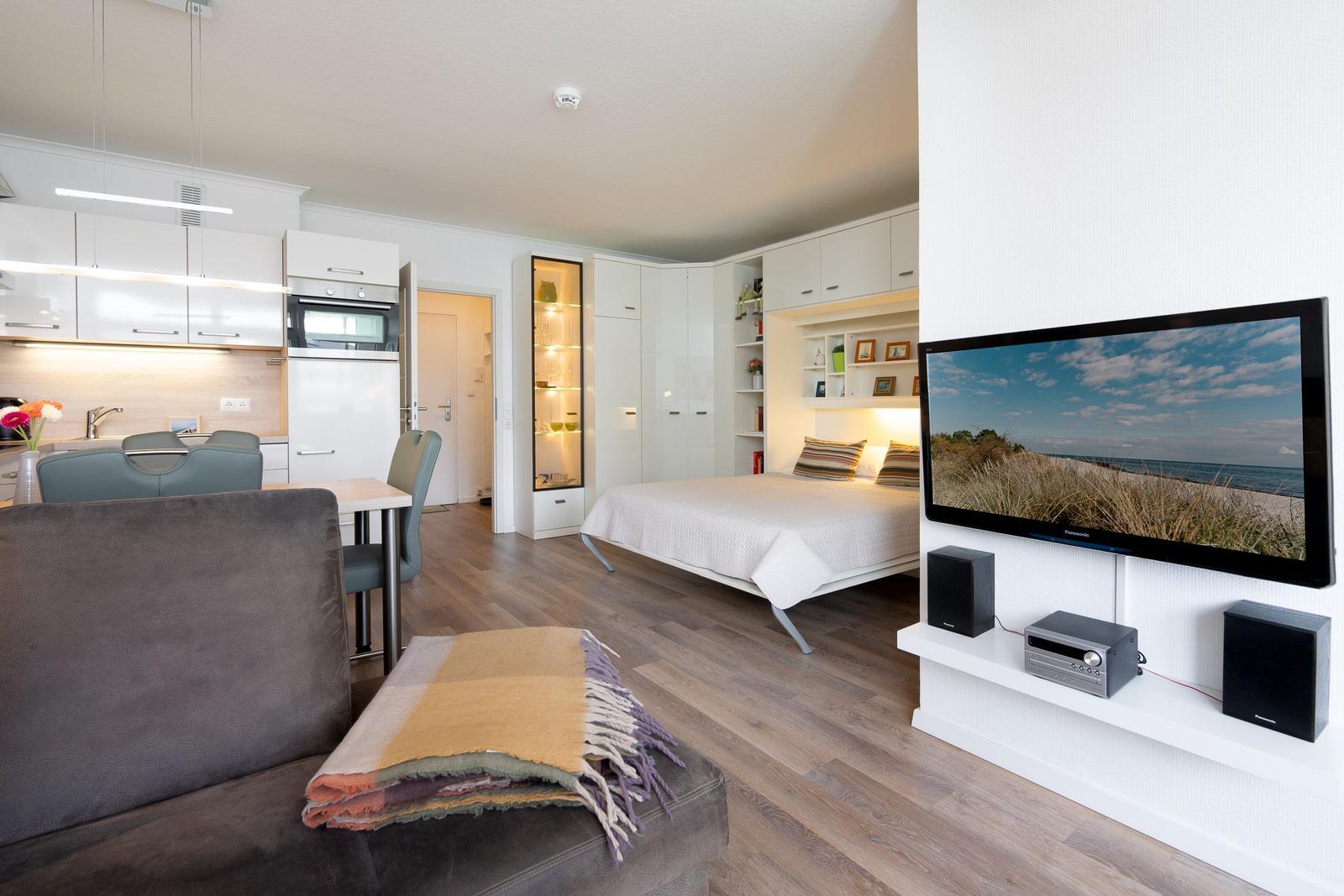 Wohnung mit Schlafzimmer, Küche und TV. Holzboden und weiße Möbel.