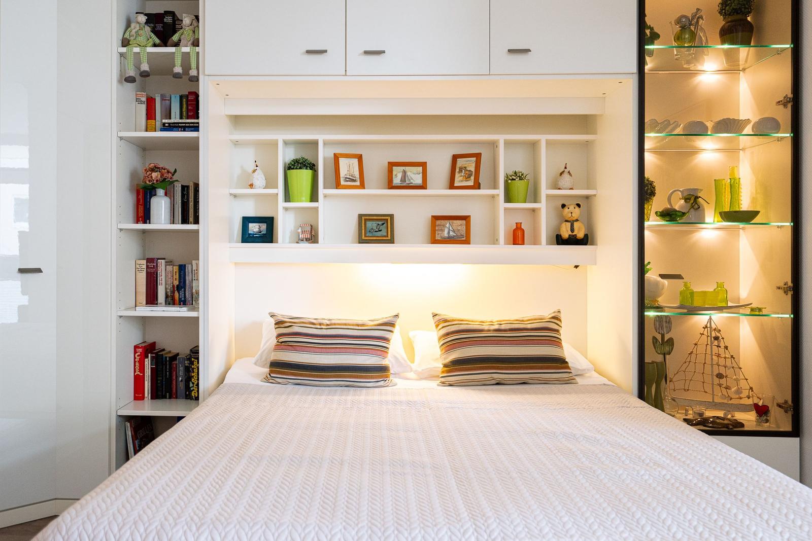 Schlafzimmer mit Bett, Regalen und beleuchteten Vitrinen.