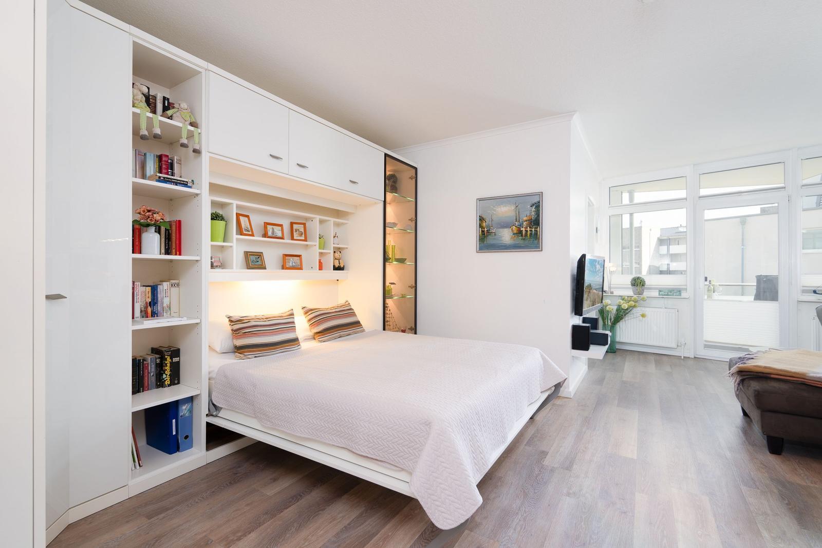 Schlafzimmer mit Bett, Bücherregal und TV. Holzboden und großes Fenster.