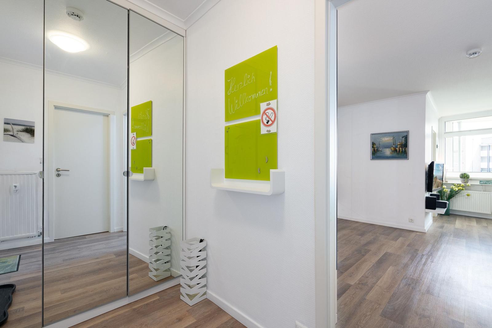 Eingangsbereich mit Spiegeltüren und grünem Willkommensschild. Holzdielen und weißer Wand.