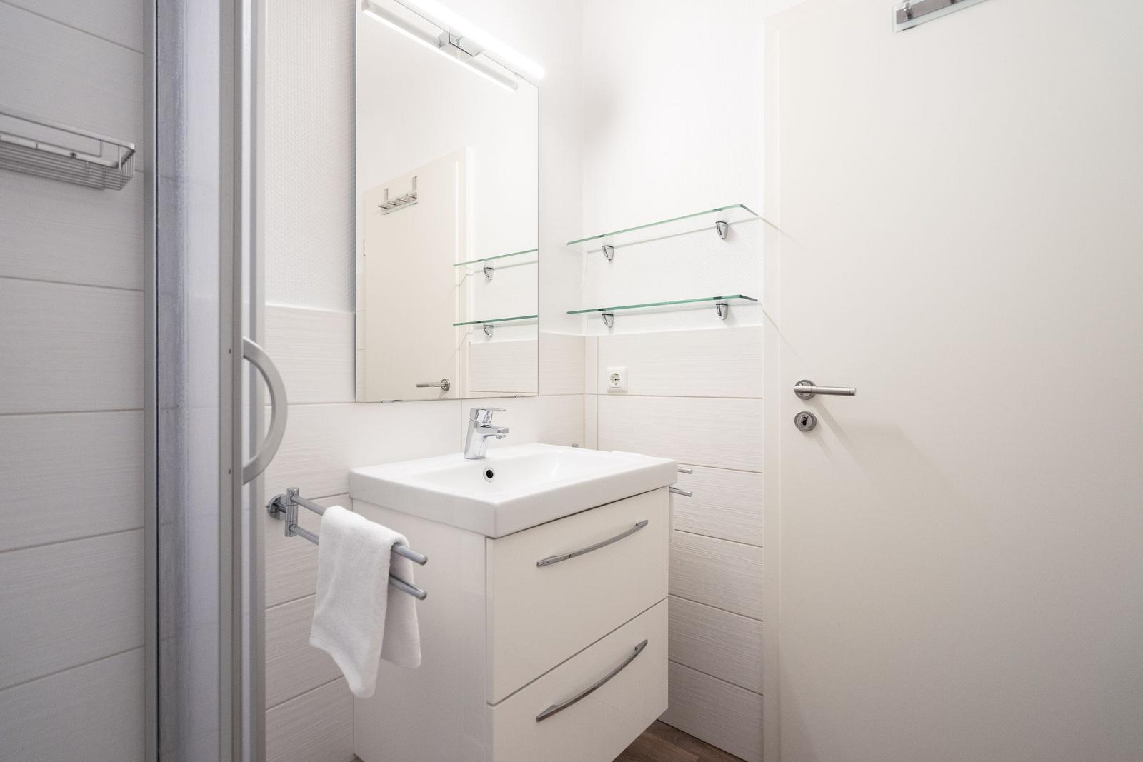 Badezimmer mit Waschbecken, Spiegel und Dusche. Glasregale und Handtuchhalter.