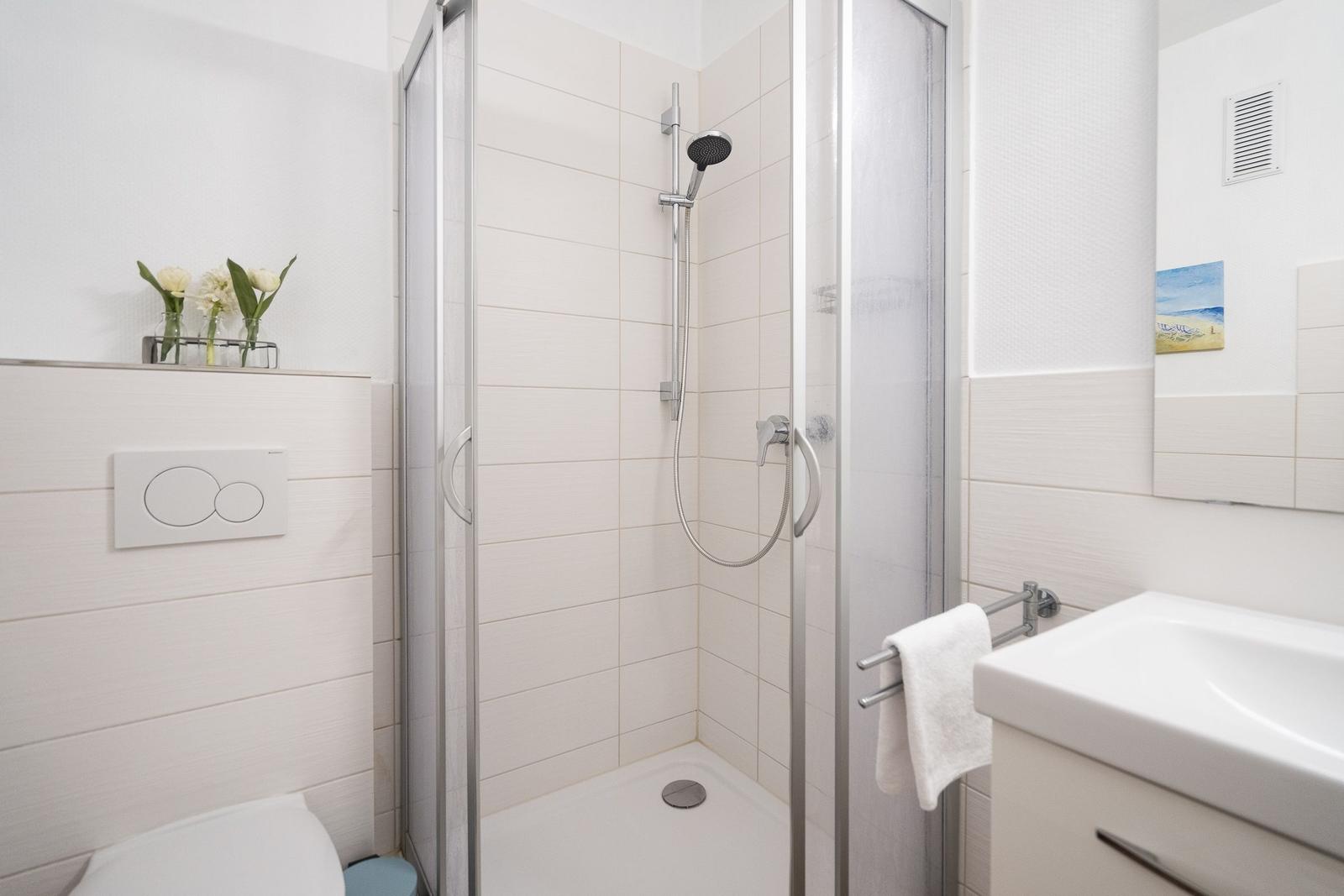 Badezimmer mit Dusche, Waschbecken und Toilette. Weiße Fliesen und moderne Einrichtung.