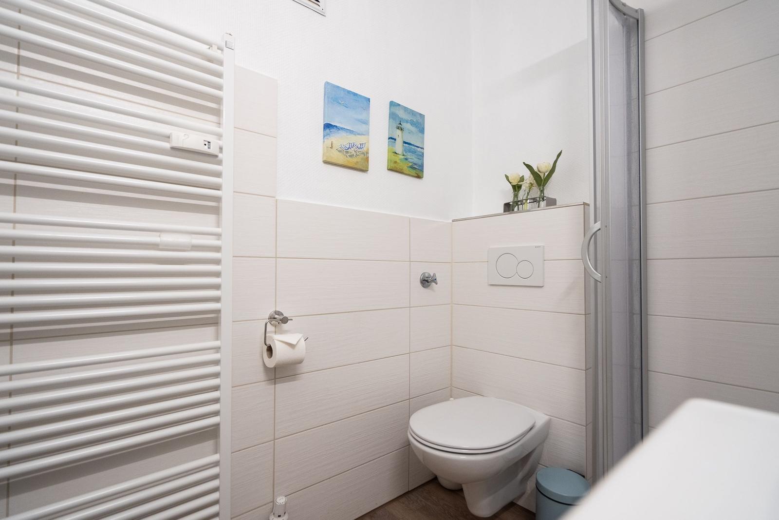Badezimmer mit Toilette, Dusche und Heizkörper. Wandbilder zeigen Strand und Leuchtturm.