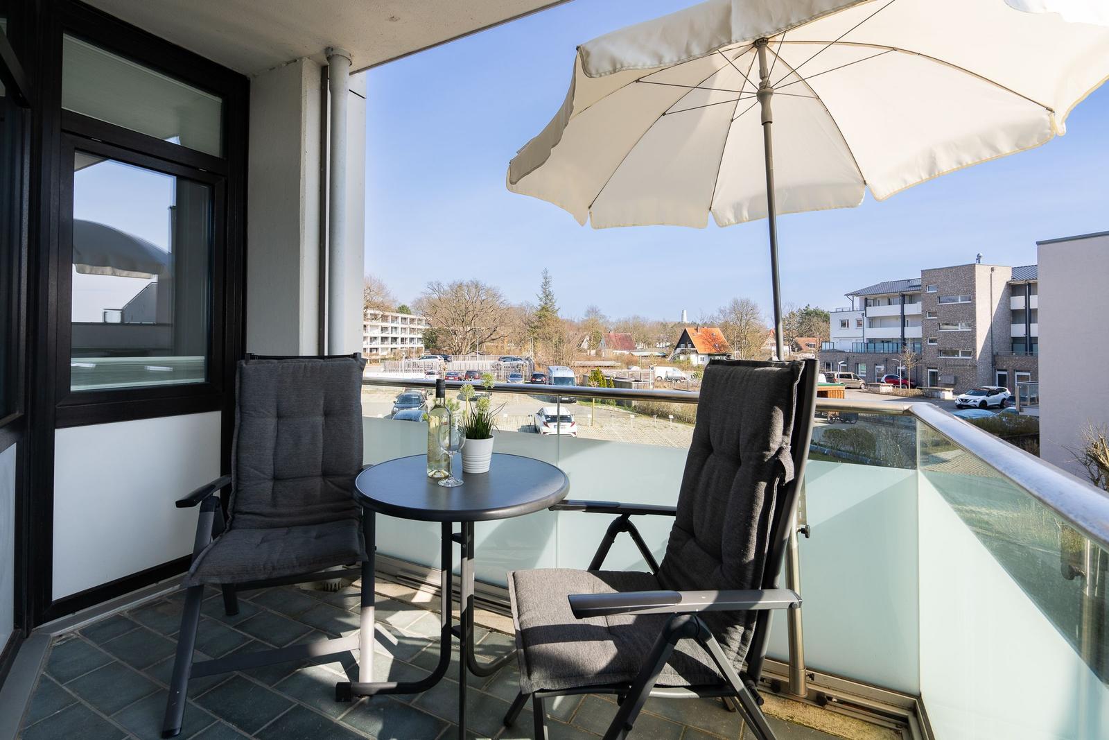 Terrasse mit Tisch, zwei Stühlen und Sonnenschirm. Glasgeländer und Blick auf Wohngebiet.