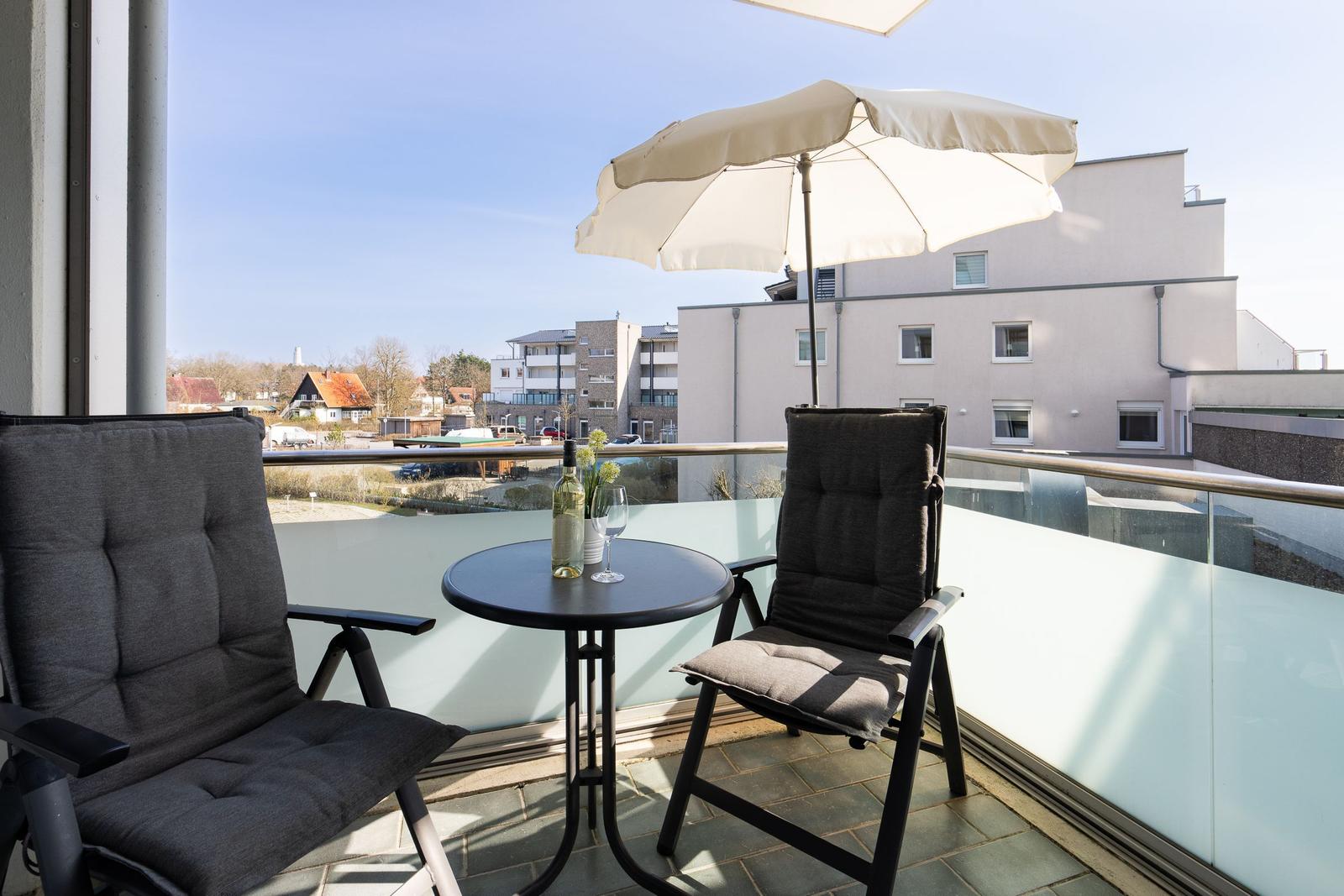 Terrasse mit zwei Stühlen, Tisch und Sonnenschirm. Blick auf Gebäude und Grünflächen.