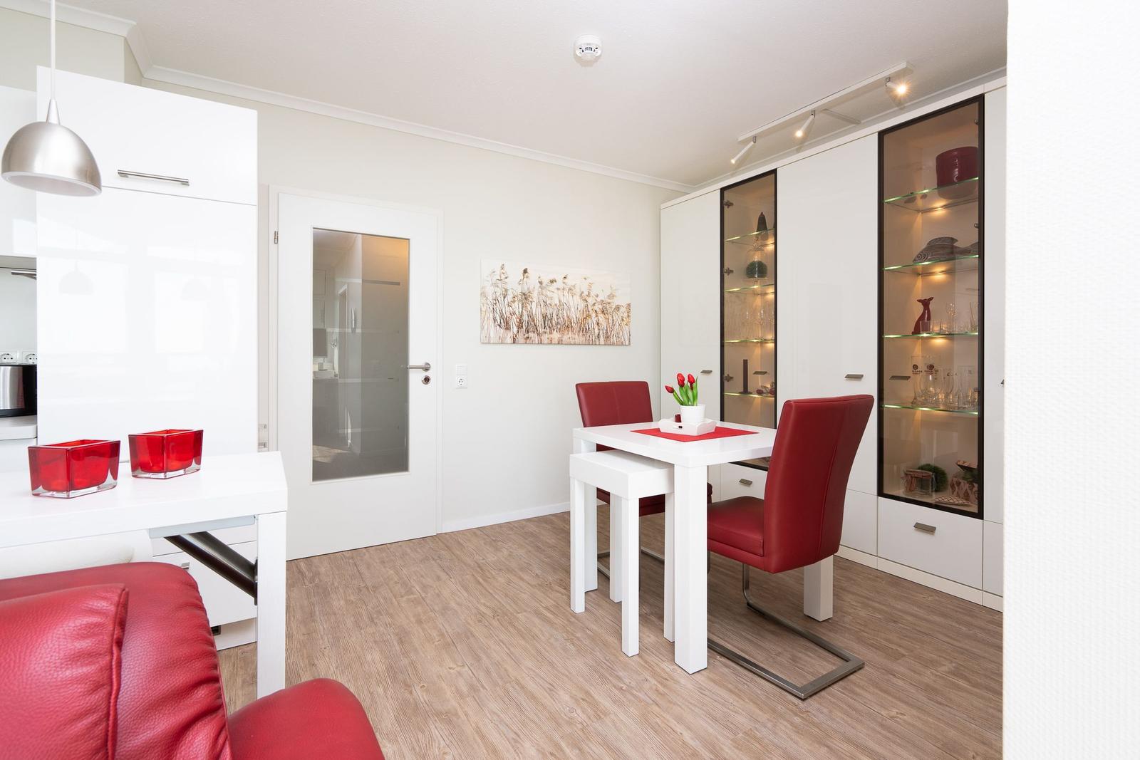 Living-dining area with white table, red chairs, and glass cabinets. Wooden floor and white walls.