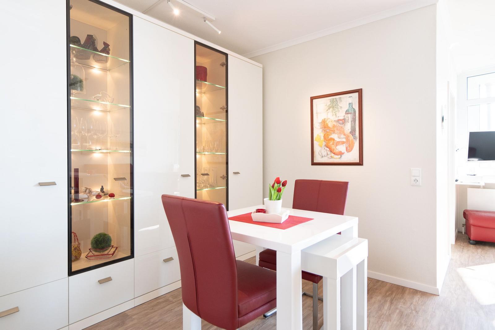 Dining room with white table, red chairs, and glass-front cabinets. Wall art and wooden floor.