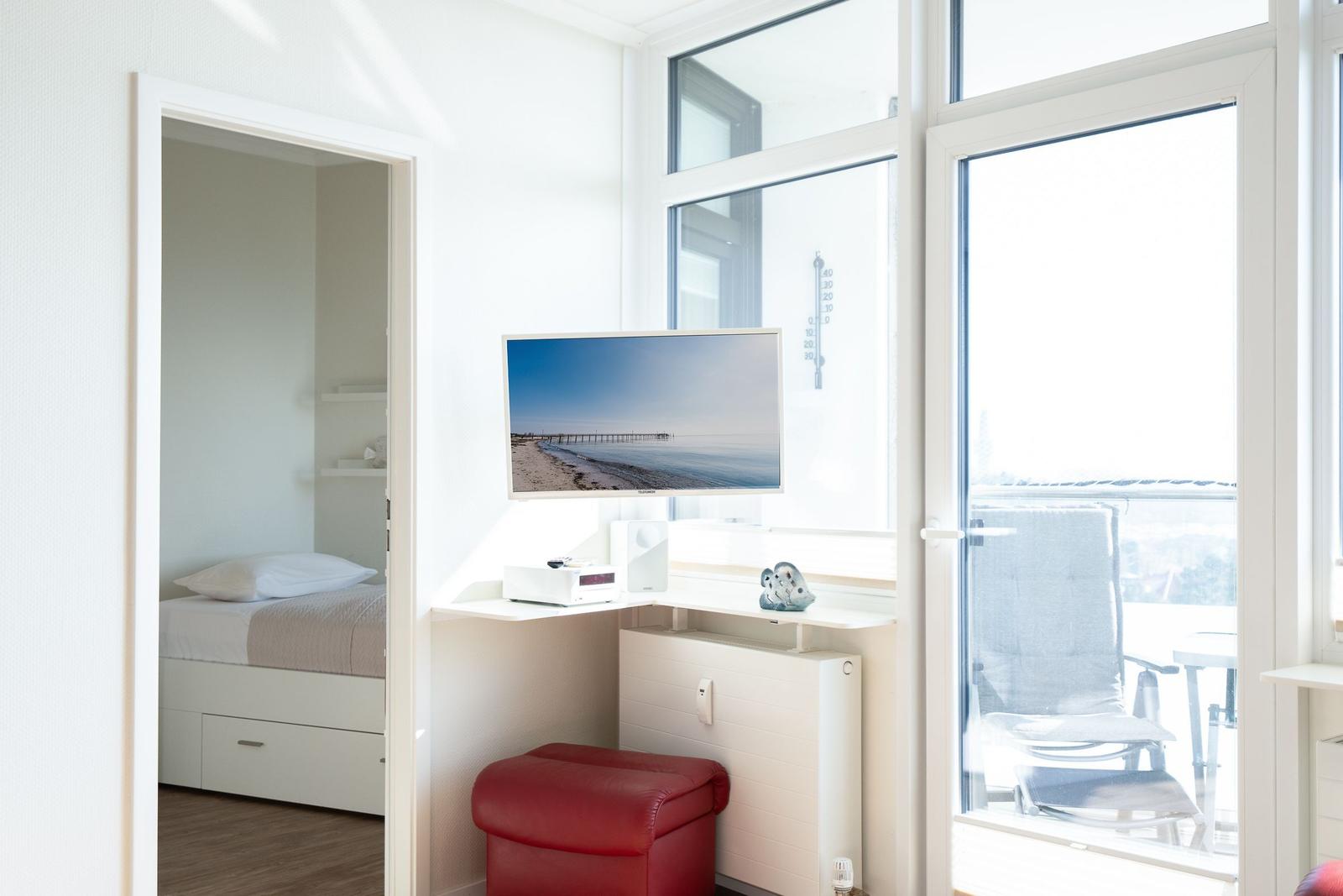 Living area with TV, red ottoman, and balcony view. Bedroom in background.